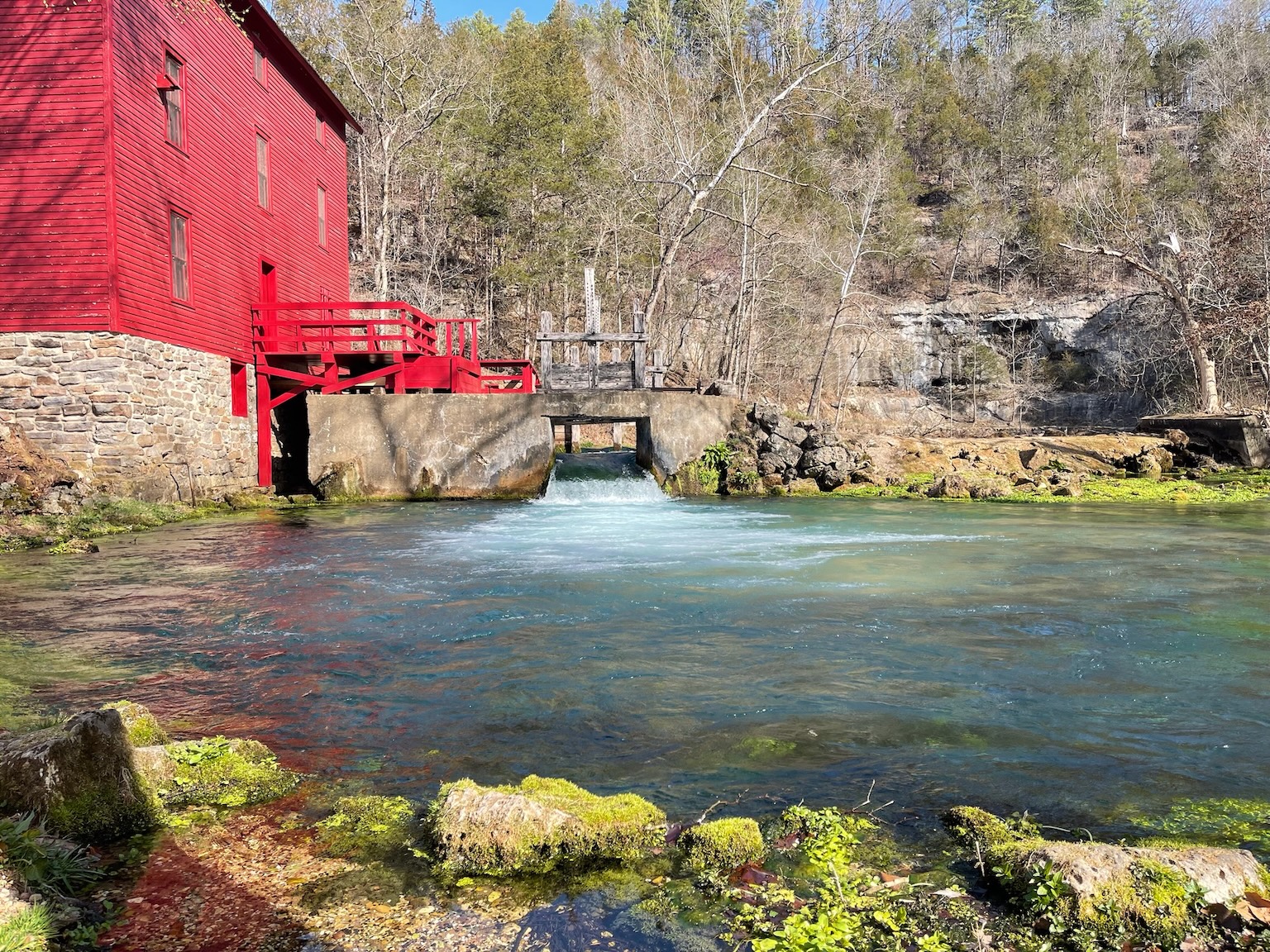 Blue water is surrounded by vibrant mossy green rocks next to the shock of red painted mill.